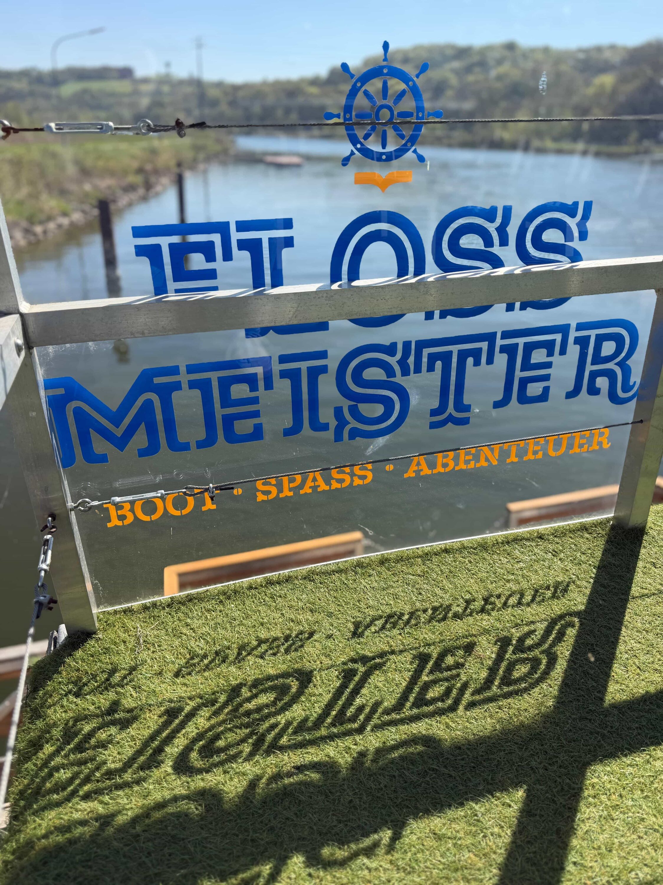 FLOSSMEISTER-Logo mit Schriftzug „Boot · Spass · Abenteuer“ auf einem Floss auf der Donau