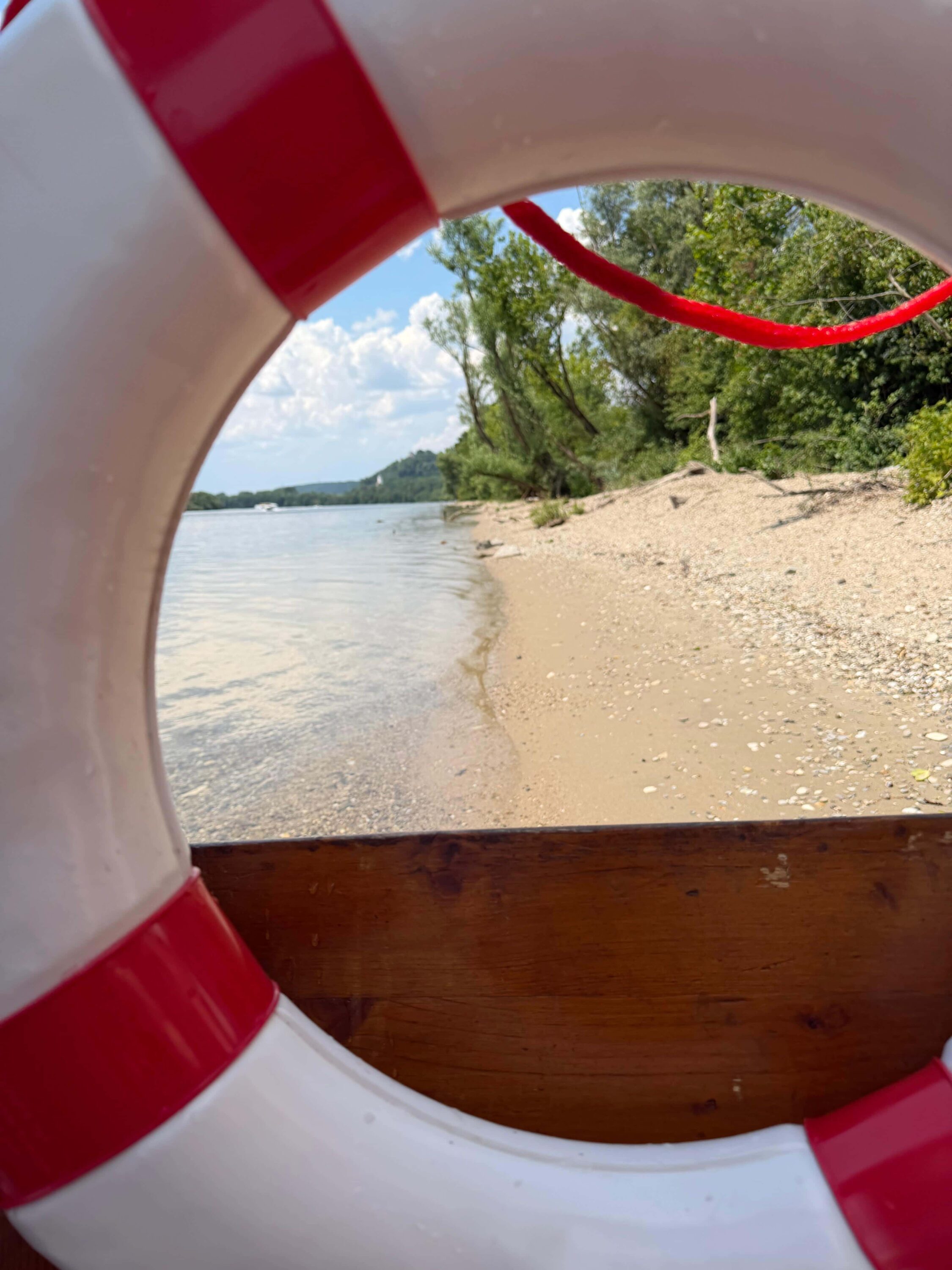 Rettungsring am FLOSSMEISTER-Floss mit Blick auf einen Kiesstrand an der Donau bei der Anlegestelle